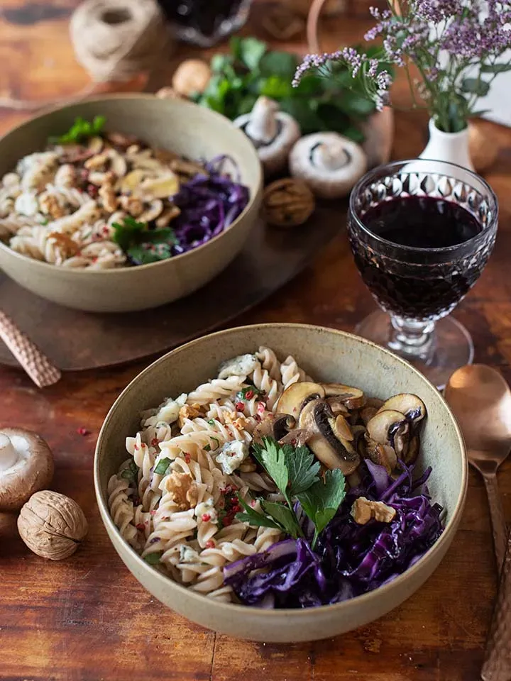 Gorgonzolanudeln mit Rotkraut, Pilzen und Walnüssen in zwei Schalen auf Holztisch mit Glas Rotwein