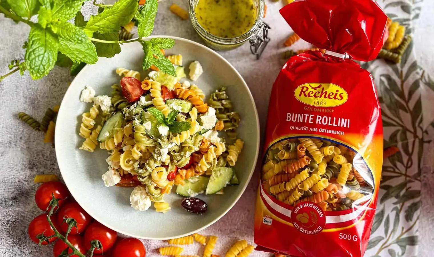 Griechischer Nudelsalat mit bunten Nudeln, Gemüse und Feta neben Packung bunter Rollini Nudeln