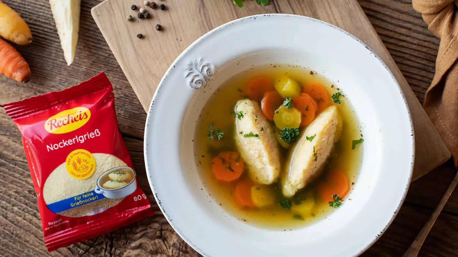 Grießnockerlsuppe: Teller mit Brühe, Gemüsescheiben und Nockerl neben Packung Nockerlgrieß auf Holzuntergrund