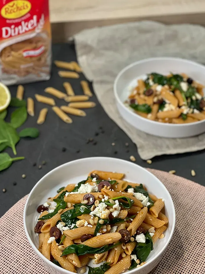 Harissa-Pasta: zwei Schalen Dinkel-Penne mit Spinat, Oliven und Feta, daneben Nudelpackung