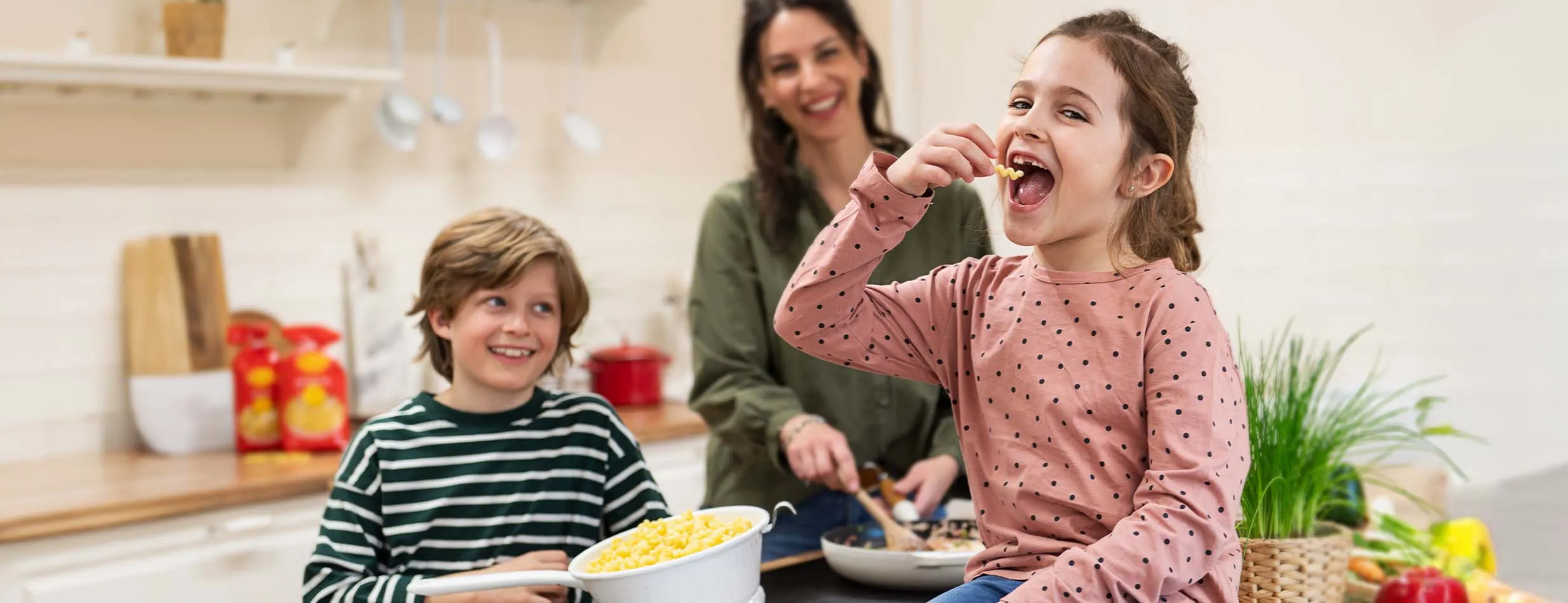 Mädchen stibitzt eine Nudel, daneben ihr Bruder und die Mutter beim Kochen in der Küche