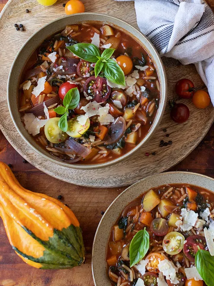 Herbst-Minestrone mit Nudelreis in zwei Schalen, daneben frische Tomaten, auf Holzuntergrund