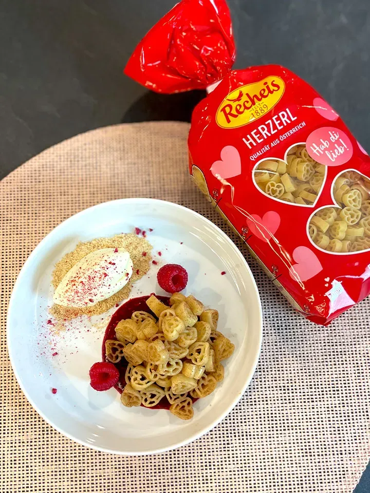 Süße Herzerlnudeln mit Himbeeren und Sahne auf Teller neben roter Packung Herznudeln