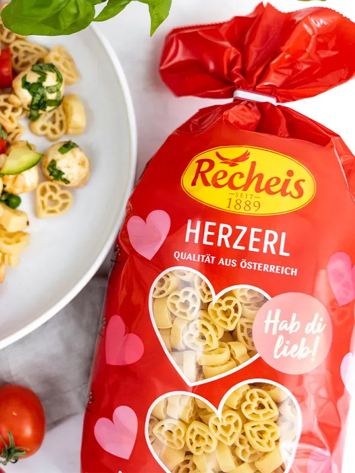 Herzerl-Caprese-Nudelsalat mit Basilikum auf Teller neben roter Packung Herzerl Nudeln