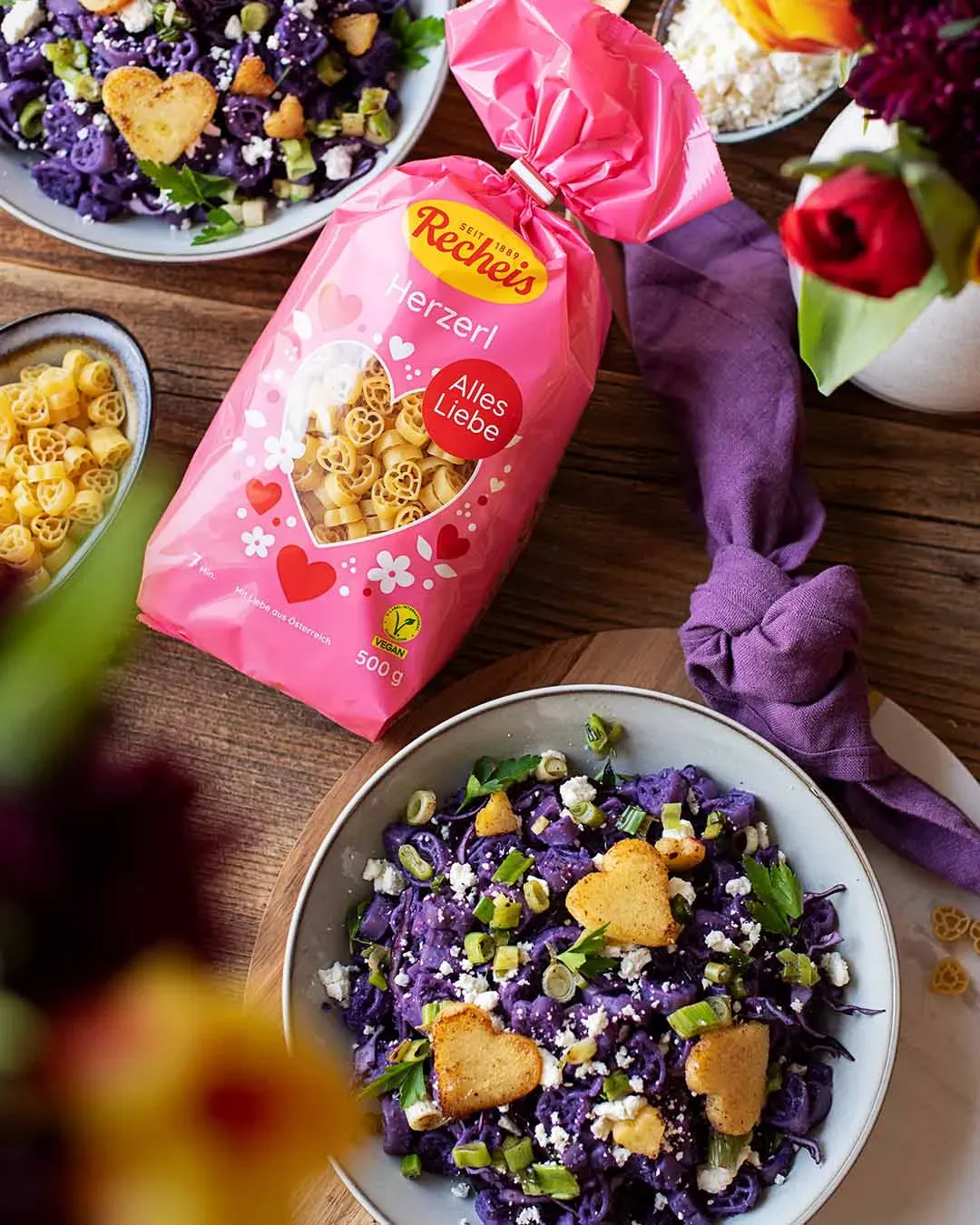 Herzerl-Nudelbowl mit Pesto: herzförmige Pasta und lila Gericht mit Kräutern und Käse auf Holztisch, dazu die Packung