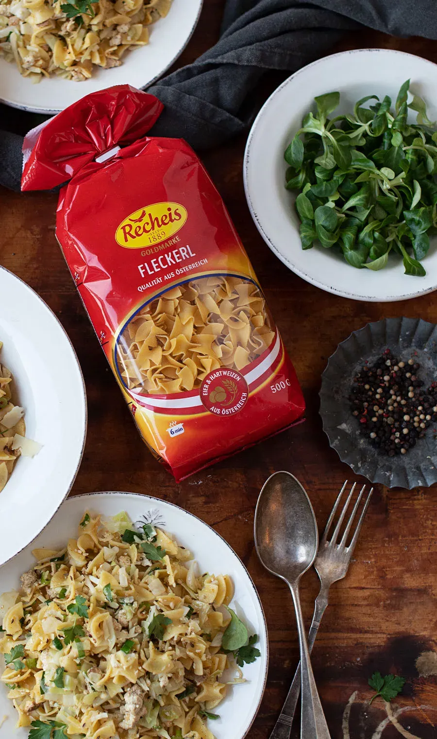 Herzhafte Krautfleckerl: Packung Fleckerl-Nudeln auf einem Tisch mit Tellern Pasta, Salat und Besteck