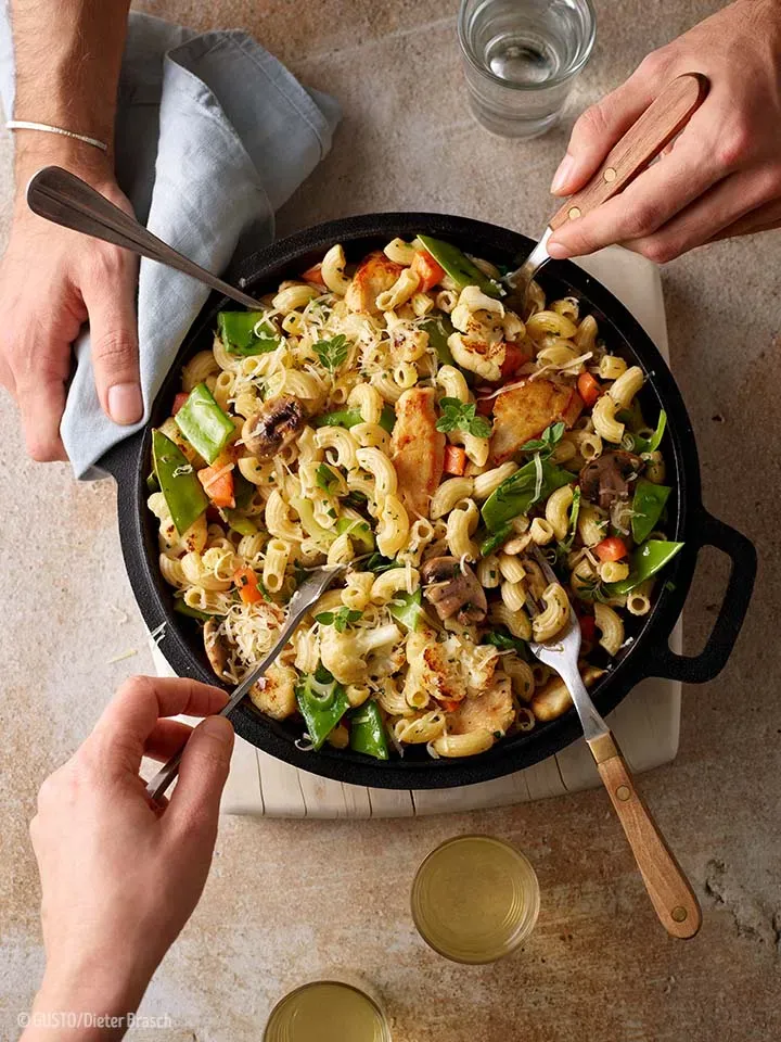 Menschen essen gemeinsam Hörnchengröstl mit Huhn und Gemüse mit Gabeln aus der Pfanne