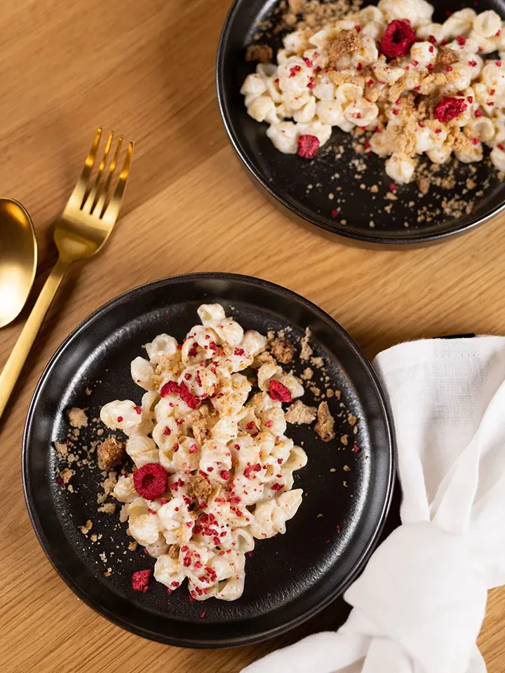 Honig-Milchnudeln mit getrockneten Himbeeren als Dessert auf zwei schwarzen Tellern auf Holztisch