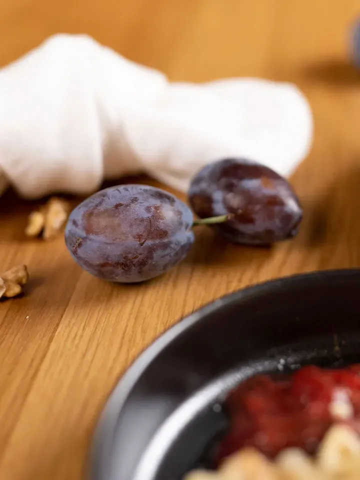 Zwei Pflaumen für Honig-Milchnudeln mit Zwetschgenröster auf Holztisch neben weißem Tuch und schwarzem Teller