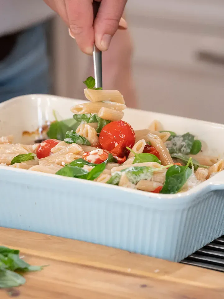 Im Ofen geschmorte Paradeiser mit Feta und Penne: Hand nimmt Gabel mit Pasta, Tomaten und Spinat aus der Auflaufform