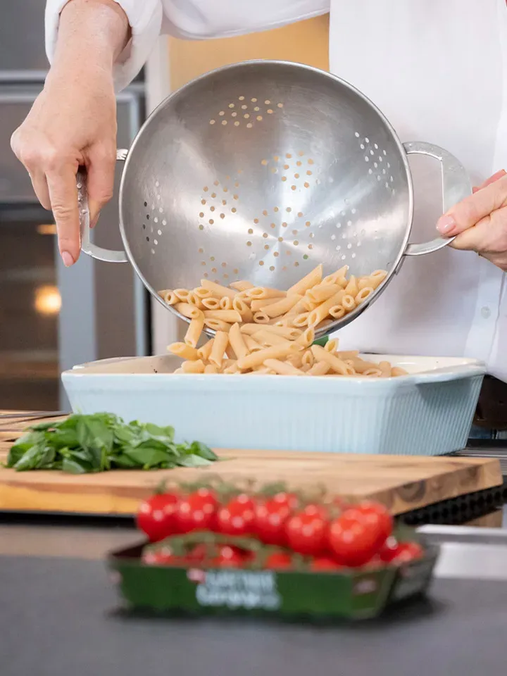 Im Ofen geschmorte Paradeiser mit Feta und Penne: Person gießt gekochte Nudeln aus dem Sieb in eine Auflaufform