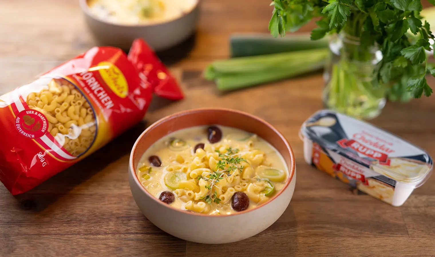 Käsesuppe mit Schmelzkäse und Nudeln in einer Schüssel, daneben Nudeln, Frischkäse und Kräuter