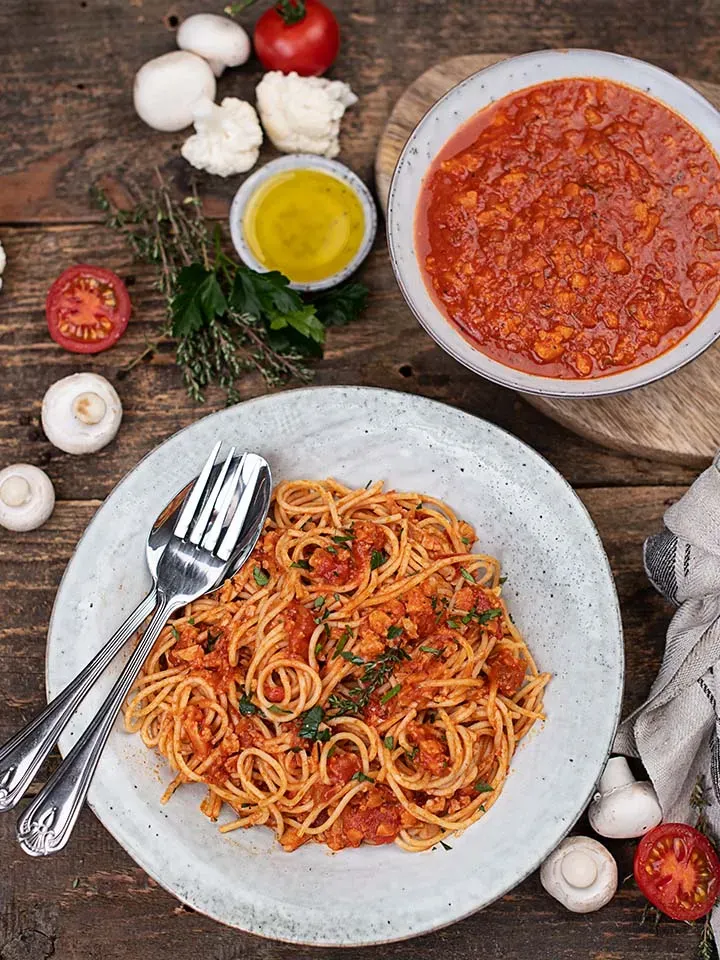 Karfiol-Bolognese mit Spaghetti in Tomatensauce auf einem Teller, dazu frische Zutaten auf Holztisch