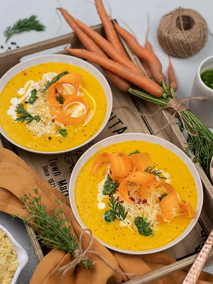 Karottencremesuppe mit Petersilienpesto in zwei Schalen mit Kräutern und Karottenröllchen auf einem Tablett
