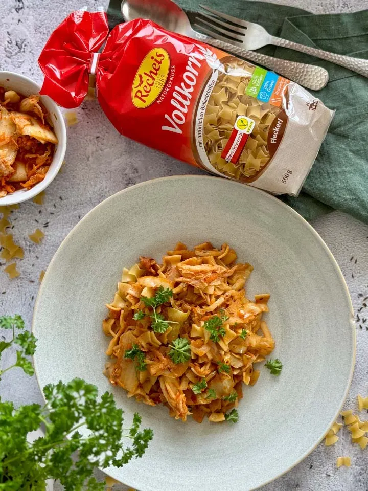 Veganes Kimchi-Fleckerl aus Vollkornnudeln mit Kräutern auf Teller neben Packung und Besteck