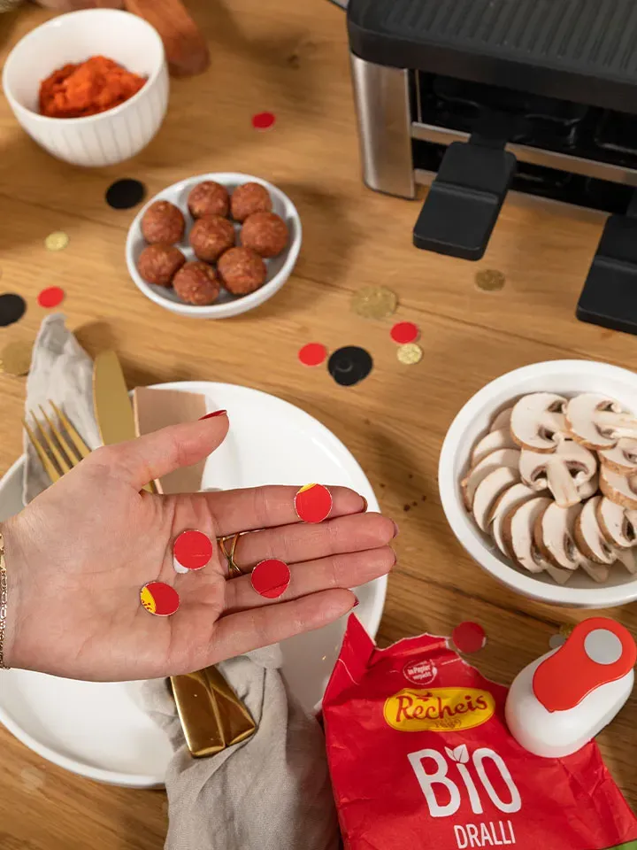 Hand hält selbstgemachtes Konfetti aus roten Papierscheiben über einem gedeckten Tisch mit Pilzen und Fleischbällchen