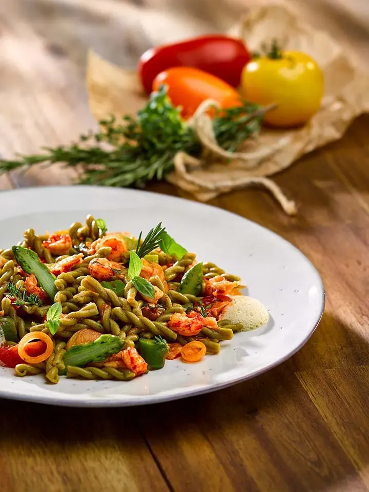 Kräuter-Treccine mit Flusskrebsen als grüner Nudelsalat auf einem Holztisch, im Hintergrund Tomaten