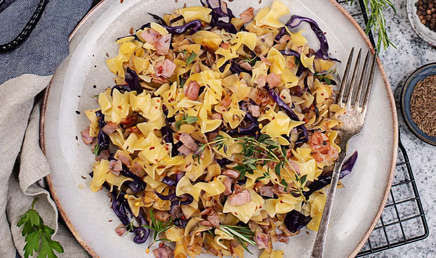 Krautfleckerl mit Rotkraut, Speck und Kräutern auf einem Teller, daneben eine Gabel
