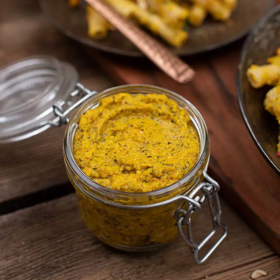 Veganes Kürbispesto in einem Glas auf Holztisch, im Hintergrund Snacks