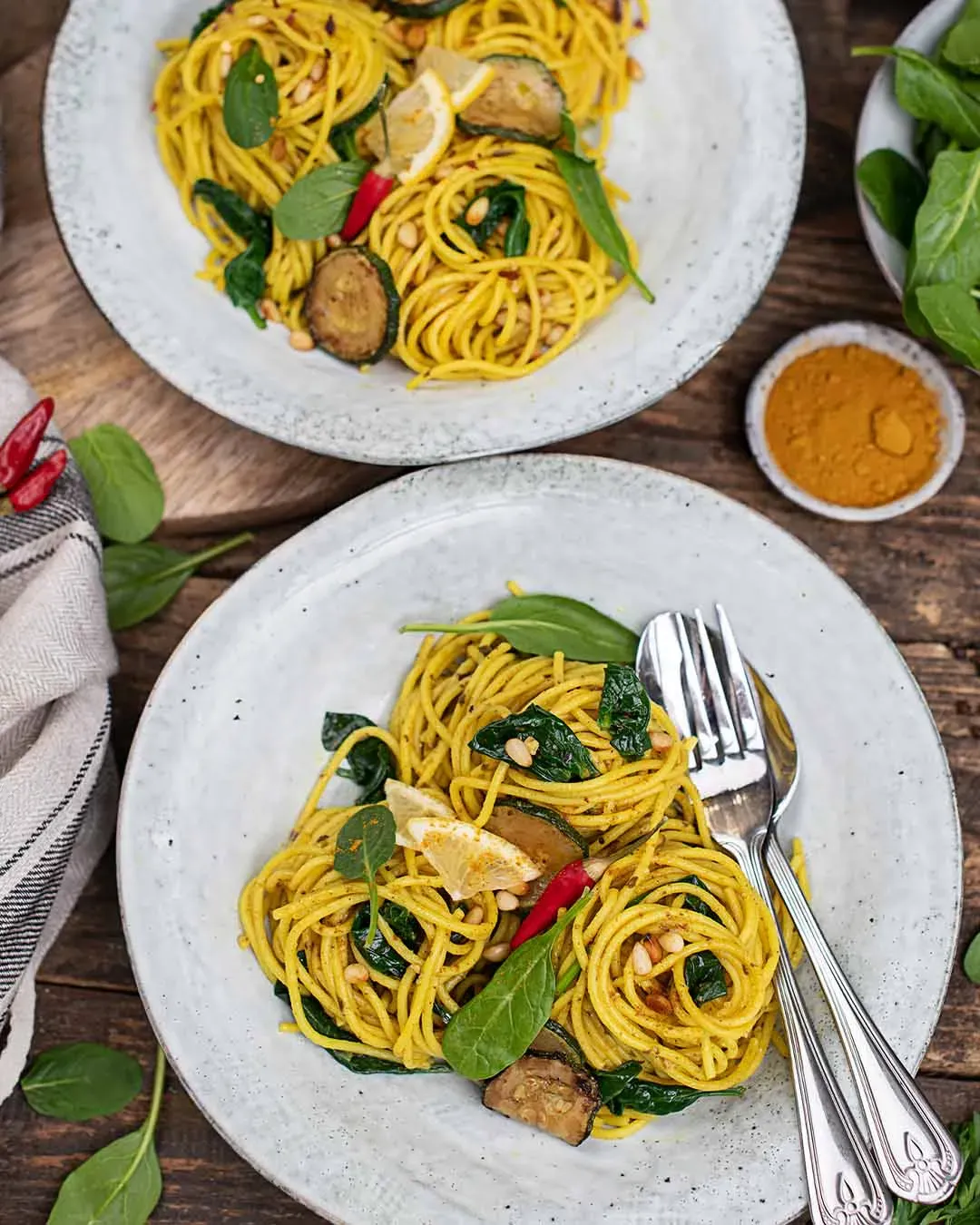 Kurkuma-Spaghetti mit Spinat und Gemüse auf zwei Tellern auf Holztisch mit Besteck