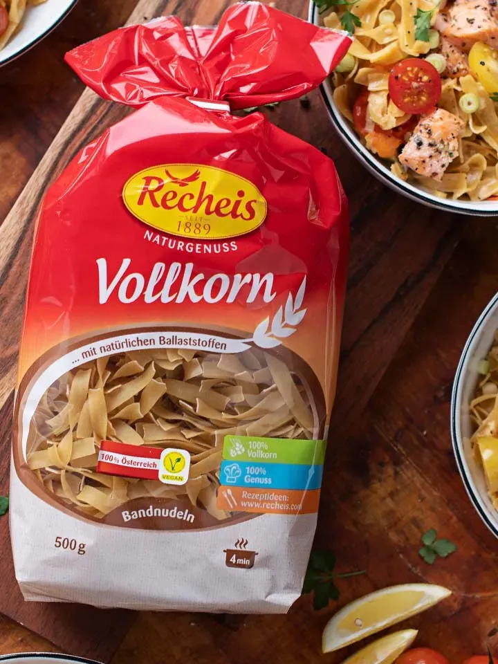 Packung Recheis Vollkorn Bandnudeln auf Holzbrett neben Lachsnudeln mit Gemüse