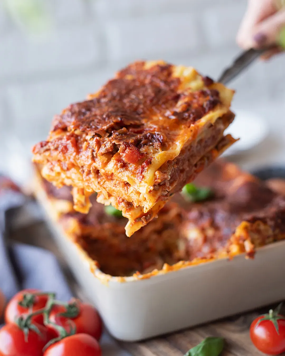 Ein Stück klassische Lasagne wird aus der Auflaufform gehoben, im Vordergrund frische Tomaten