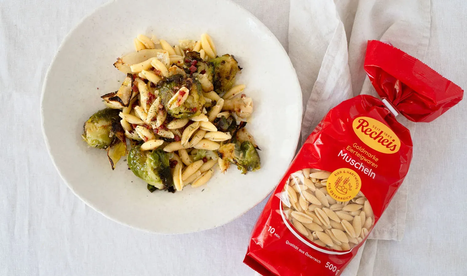 Lauwarmer Nudelsalat mit Kohlsprossen und Speckwürfeln neben einer Packung Muschelnudeln auf weißem Tuch