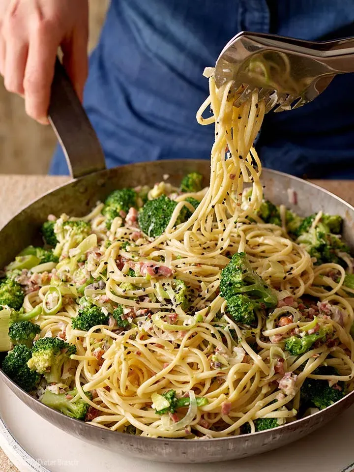 Linguine mit weißem Pesto, Brokkoli und Speck werden in der Pfanne mit Gabel und Zange angerichtet