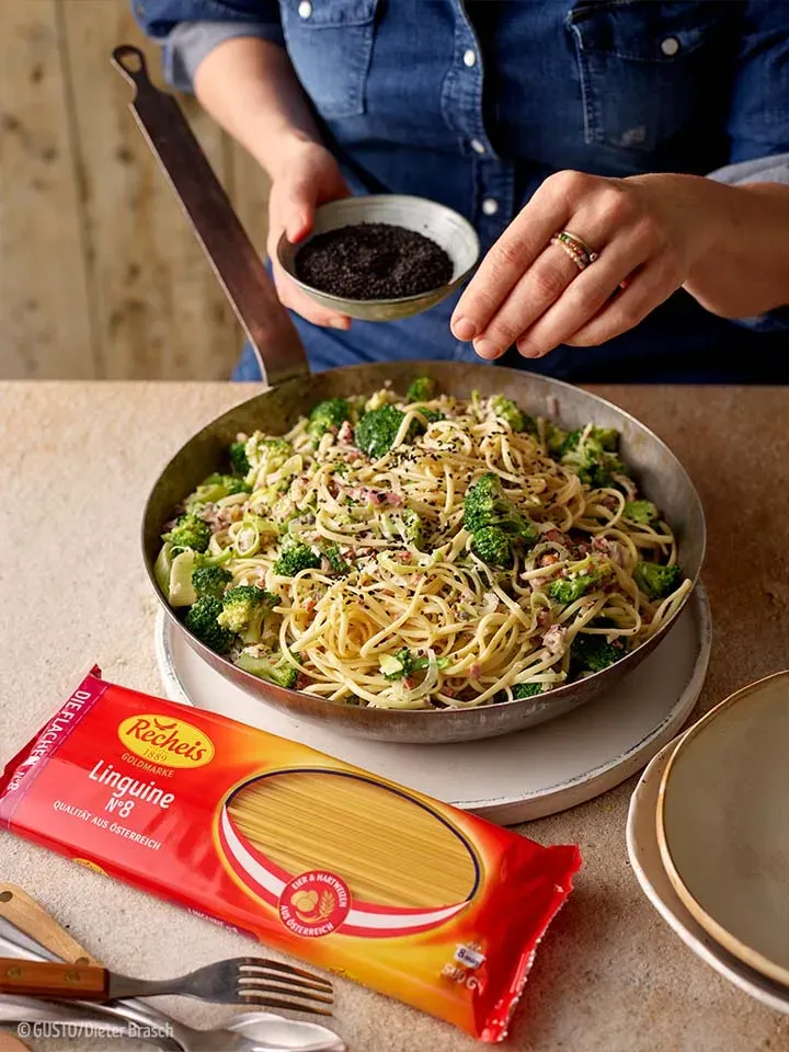 Person streut schwarzen Sesam über Linguine mit weißem Pesto und Brokkoli in der Pfanne