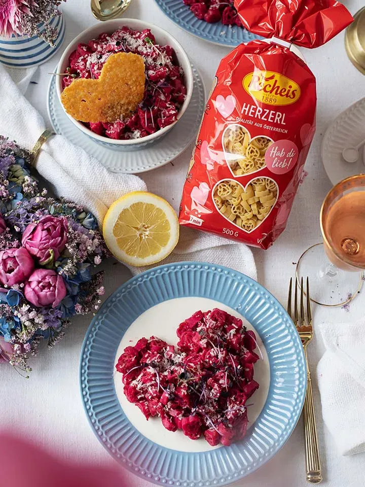 Mac and Cheese mit Roter Bete: herzförmige Pasta mit roter Soße und Parmesan auf Tellern neben der Verpackung