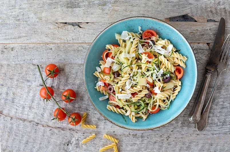 Mediterraner Low-Carb-Nudelsalat mit Tomaten und Käse auf blauem Teller mit Besteck