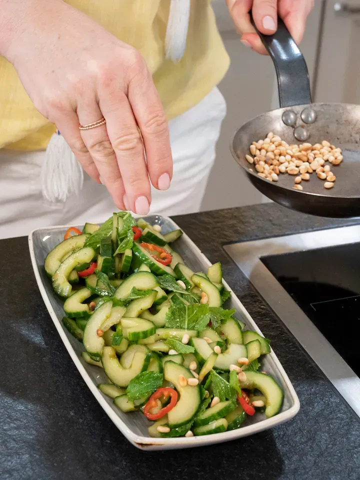Melanzani-Lasagne von Martina Hohenlohe: Hand streut geröstete Pinienkerne auf Gurkensalat mit Chili und Minze