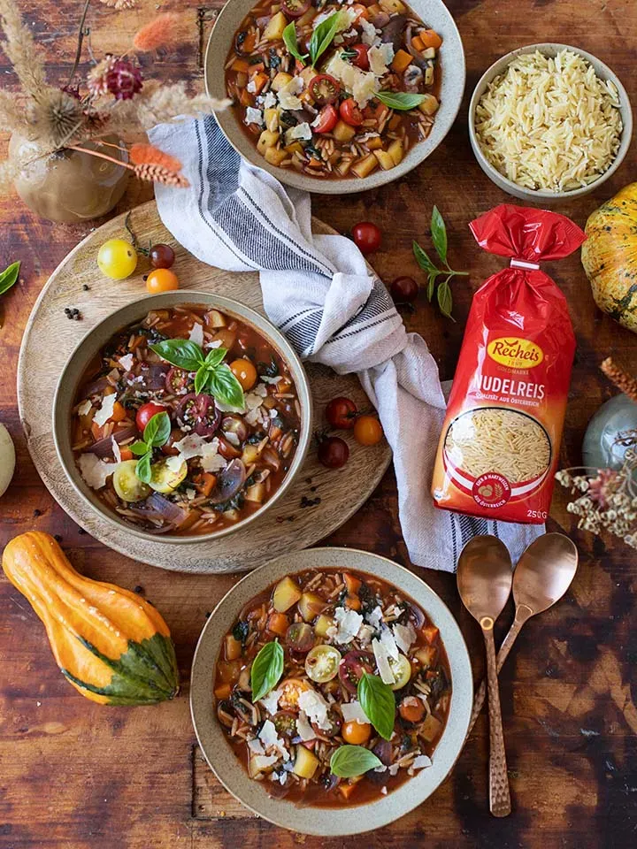 Minestrone mit Goldmarke Nudelreis und frischen Kräutern in drei Schalen auf Holztisch mit Kürbis
