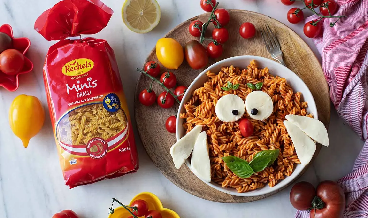 Mini-Nudeln mit Mozzarella als Eulengesicht auf einem Teller neben Nudelpäckchen und Tomaten