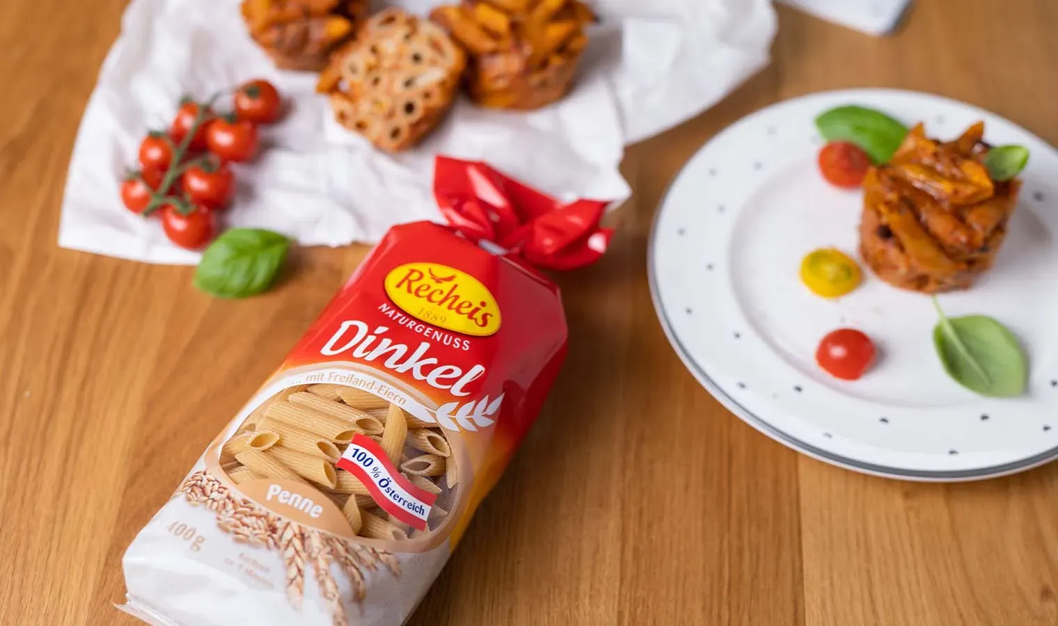Nudelmuffins mit Tomate und Mozzarella, daneben Packung Dinkel-Penne, Tomaten und Basilikum auf Holztisch