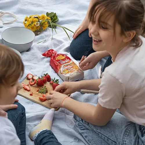 Muttertagsrezept Nudelsalat: Kinder schneiden bei einem Picknick auf einer Decke Erdbeeren auf einem Brett