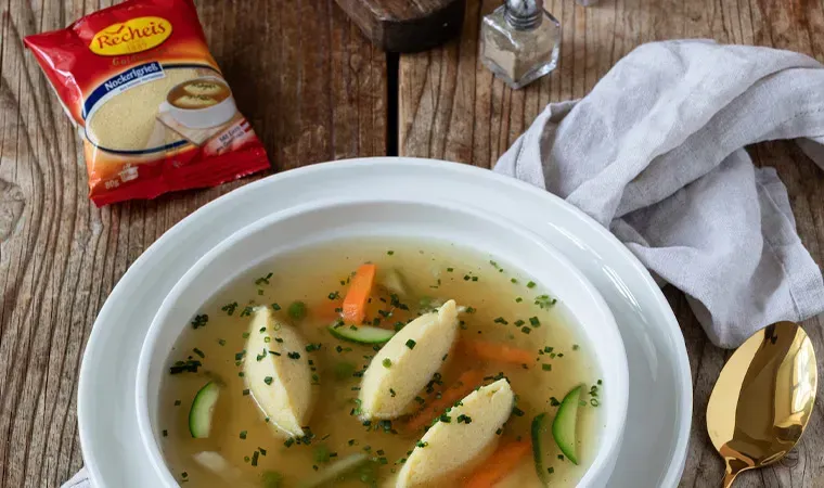 Klare Suppe mit Grießnockerln und Gemüse im tiefen Teller, daneben eine Packung Recheis Nockerlgrieß