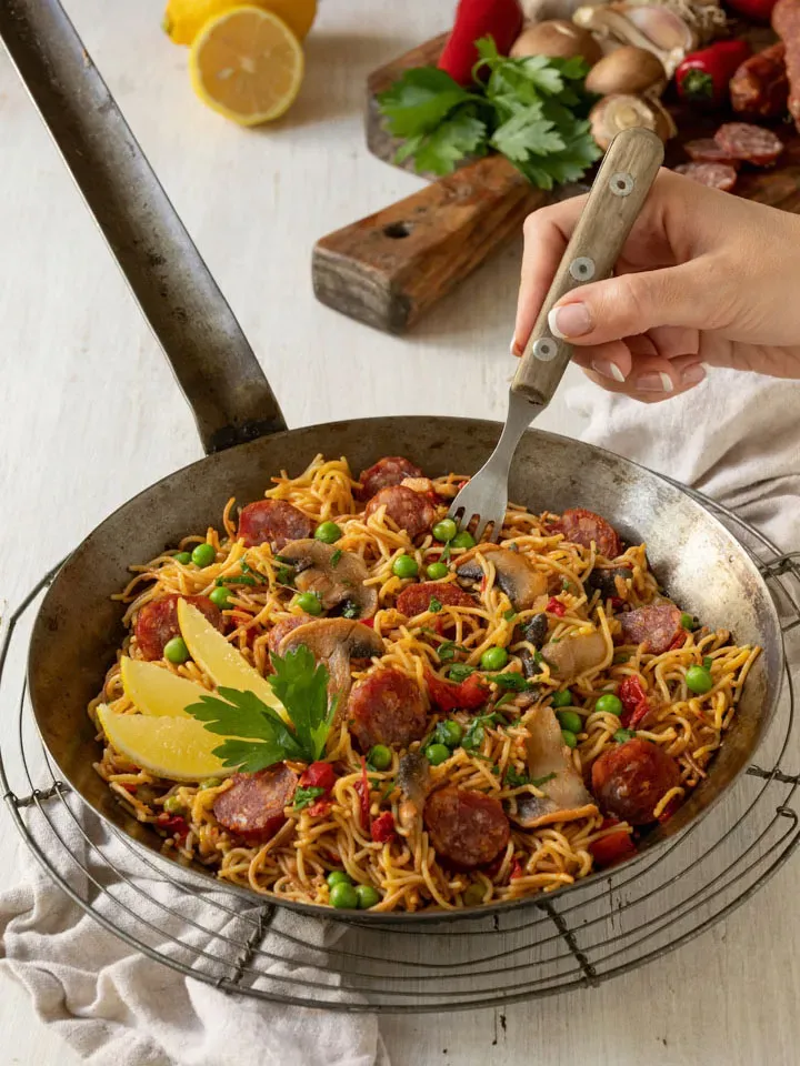 Nudel-Paella mit Chorizo, Erbsen und Pilzen in der Pfanne, Hand mit Gabel