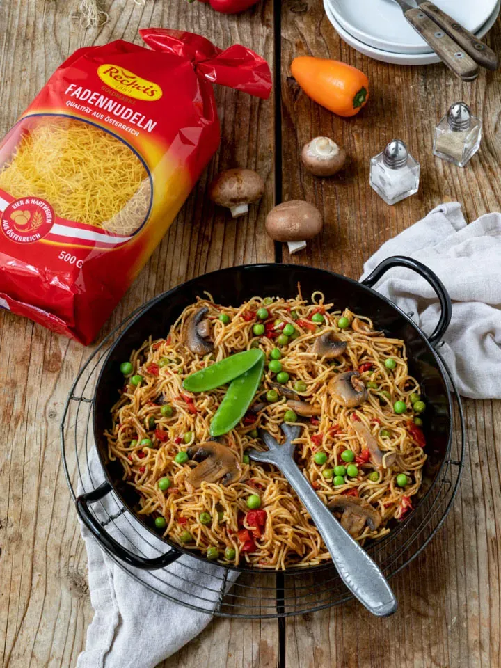 Vegetarische Nudel-Paella mit Fadennudeln, Erbsen und Pilzen in der Pfanne auf Holztisch neben Nudeltüte