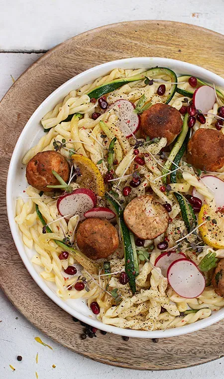 Nudel-Zucchinipfanne mit Lachsbällchen, dazu Radieschen und Granatapfelkerne, auf einem Teller