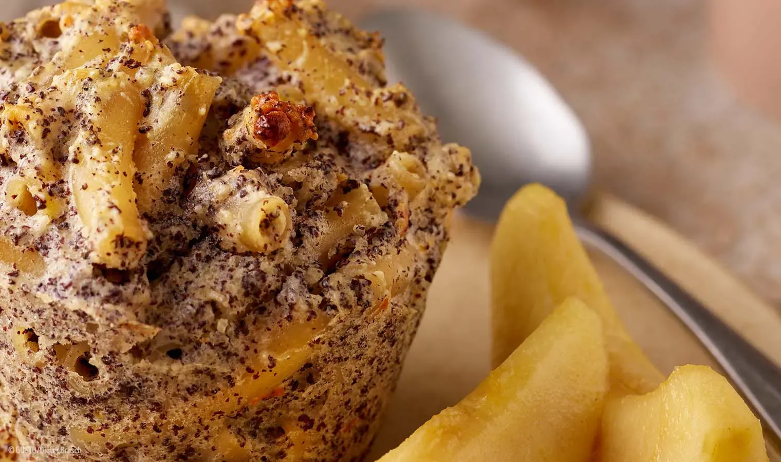 Nahaufnahme eines gebackenen Nudel-Mohnmuffins mit Apfelstücken, im Hintergrund ein Löffel