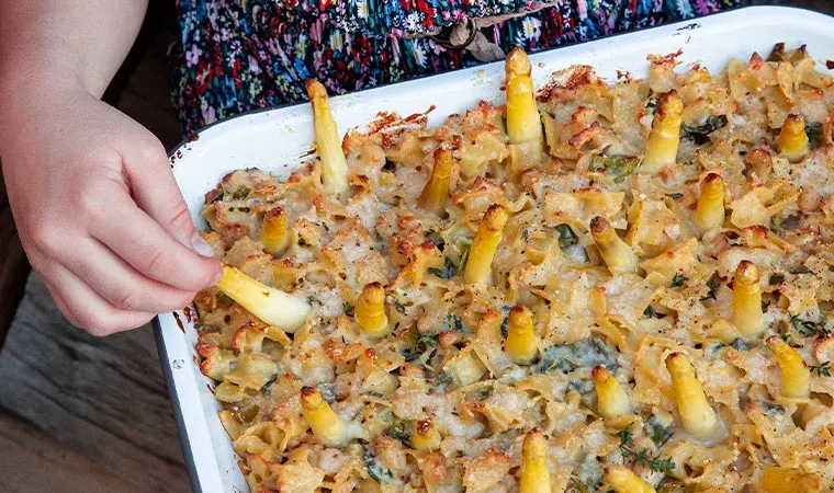 Hand nimmt weißen Spargel aus gebackenem Nudelauflauf in der Auflaufform