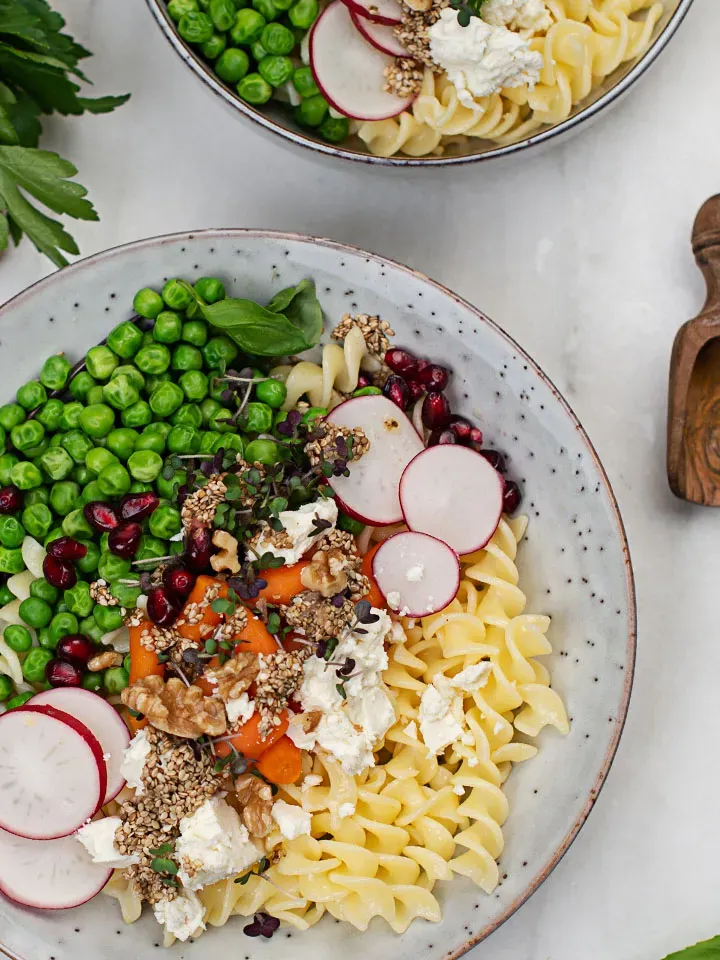 Glutenfreie Nudelbowl mit Erbsen, Radieschen, Karotten, Granatapfelkernen und Feta auf einem Teller