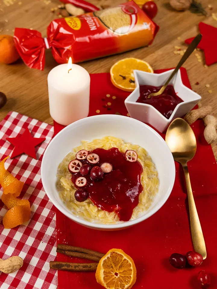 Nudelmilchreis (Orzo-Pudding) mit roter Sauce auf einem weißen Teller, dazu Kerze und Gewürze auf rotem Tuch