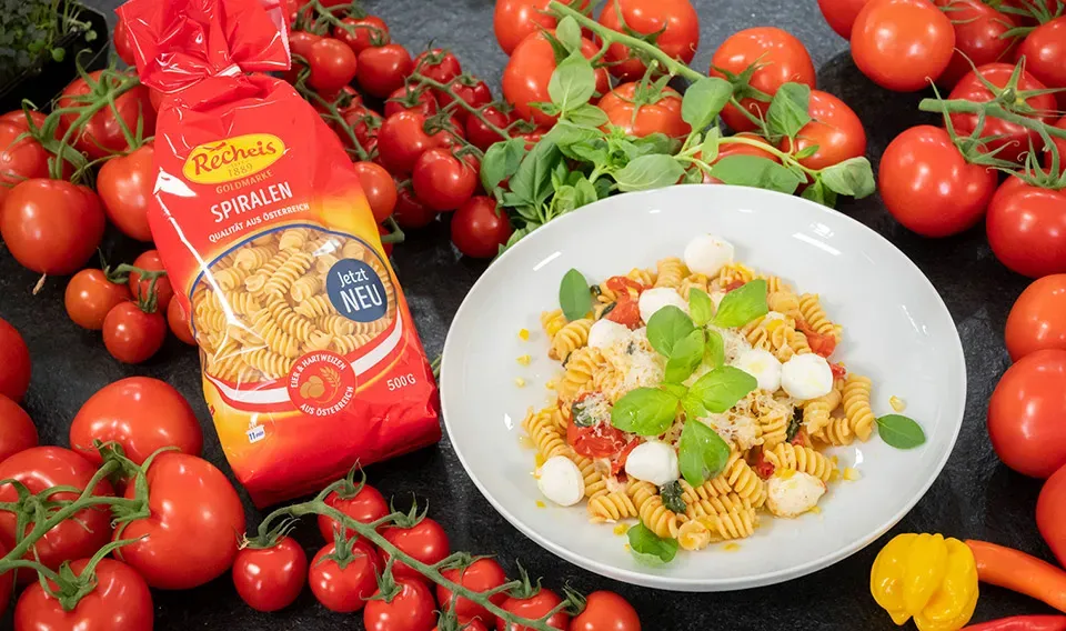 Nudeln Caprese mit Tomaten, Mozzarella und Basilikum auf Teller neben Packung Recheis Goldmarke Spiralen
