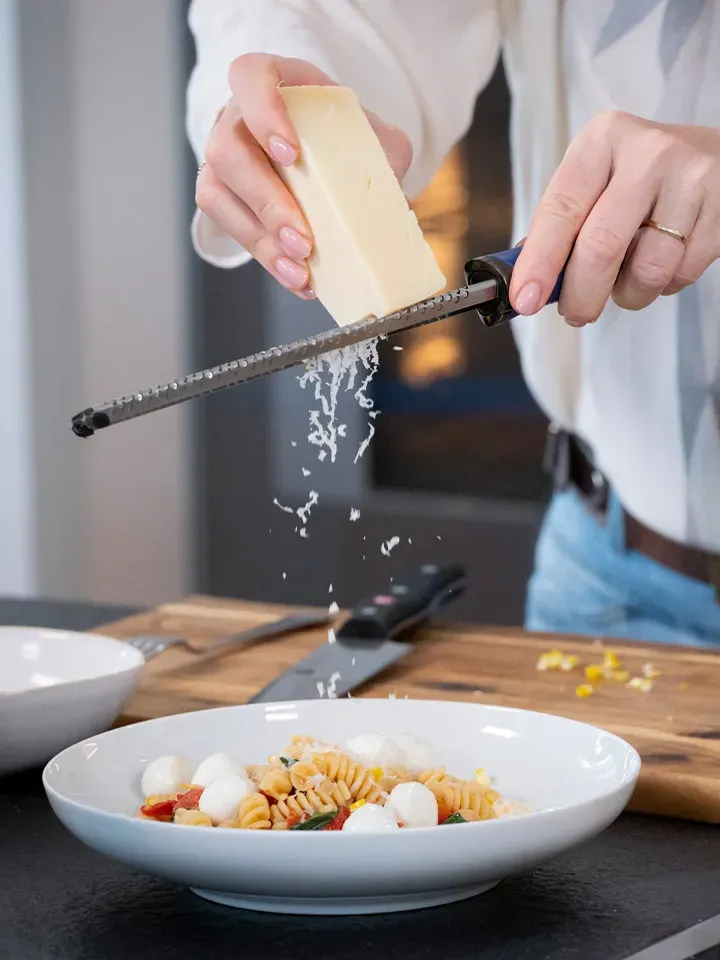 Person reibt Käse über einen Teller Nudeln Caprese mit Mozzarella