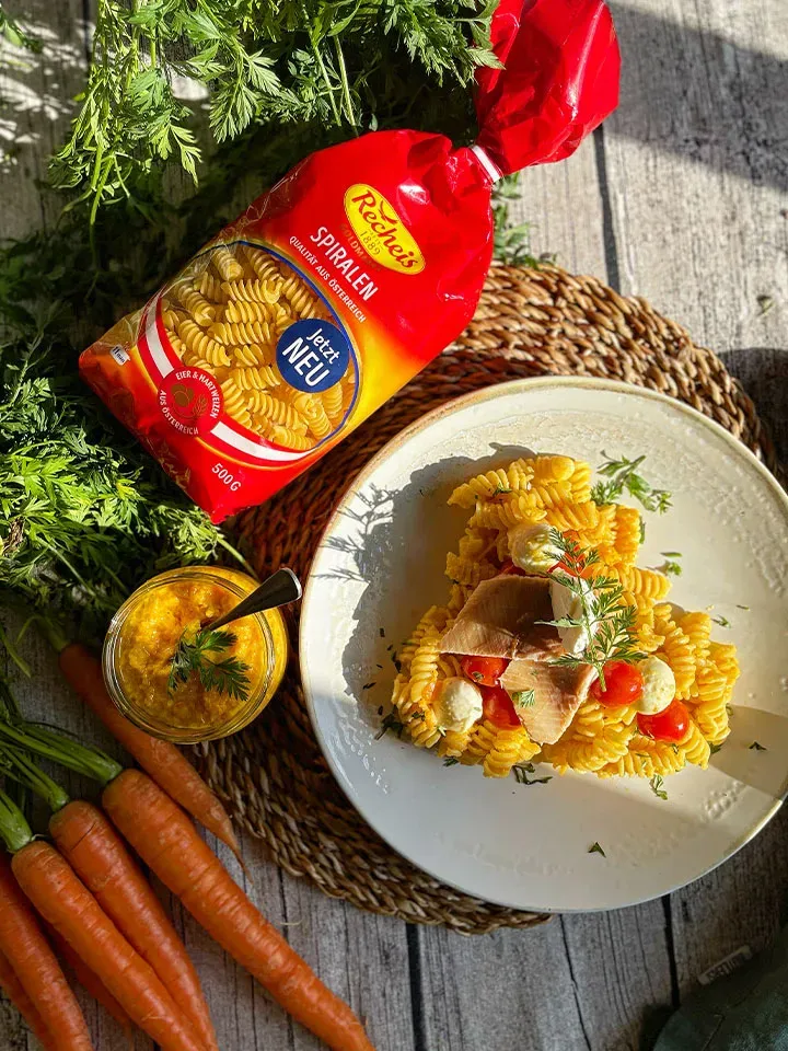 Spiralnudeln mit Karottenpesto, Schinken und Tomaten auf einem Teller neben Packung und frischen Karotten