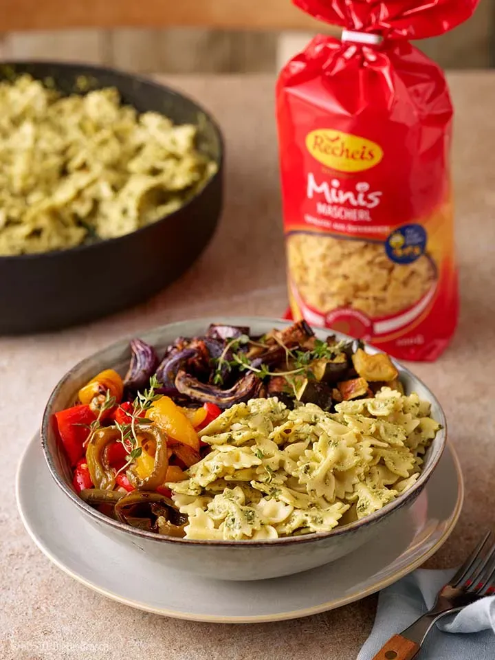 Nudeln mit Ofengemüse: Schale Farfalle mit geröstetem Gemüse und Pesto vor roter Nudeltüte