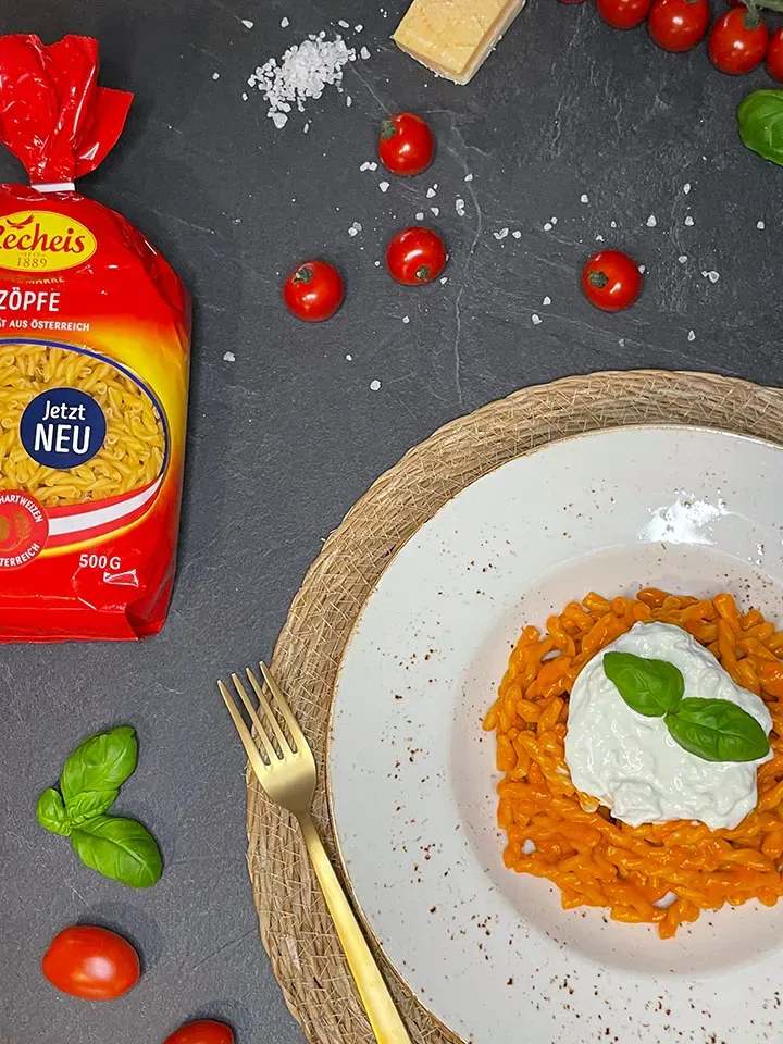 Nudeln mit selbstgemachter Datteltomatensauce, Frischkäse und Basilikum auf einem Teller, daneben Tomaten, Parmesan und Nudelpäckchen