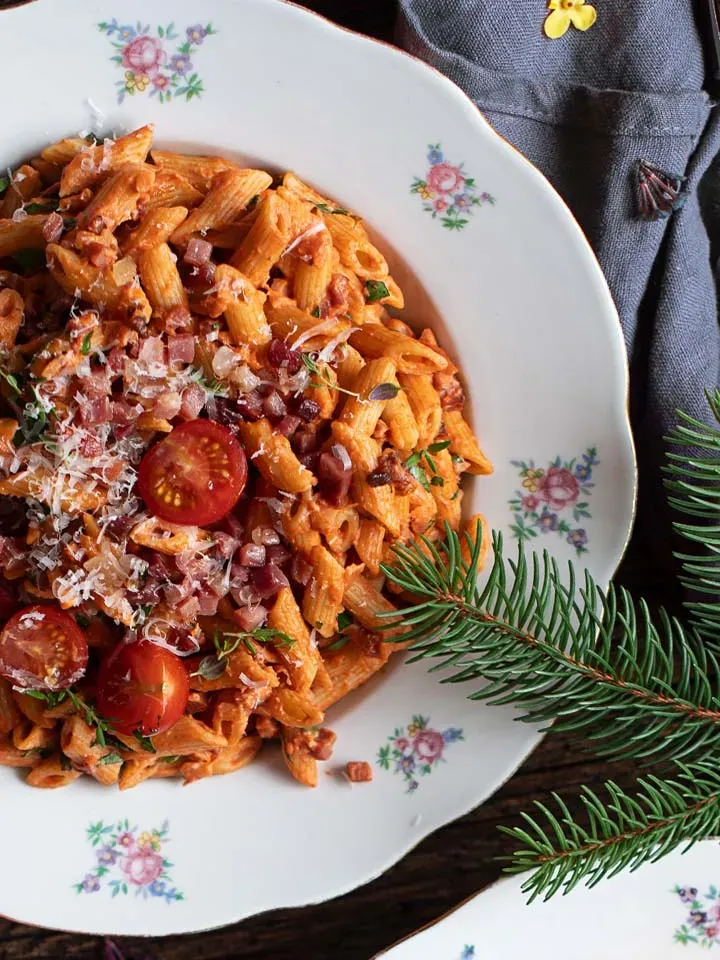 Penne mit Tomaten-Sahne-Speck-Sauce, garniert mit Kirschtomaten und Kräutern, auf einem Teller